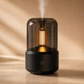 Black diffuser with smoked glass dome, warm amber glow, and soft cool mist on a neutral beige surface.